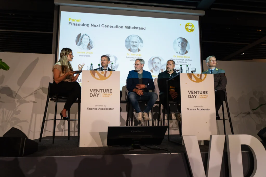 Moderatorin für Wirtschaft und Finanzen Janine Utsch auf einem Hocker sitzend neben vier Experten bei der Moderation eines Panels auf dem Finance Summit Stuttgart